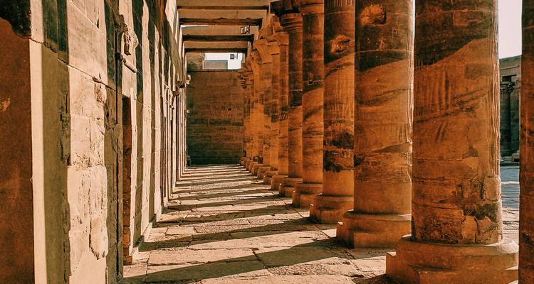 Sunlit sandstone colonnade of an ancient Egyptian temple with repeating shadows.