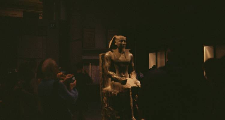 Dimly lit museum hall with blurred statue in center and visitors taking photos around it.