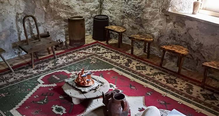 Intérieur traditionnel albanais rustique avec tapis à motifs, petits tabourets en bois et poterie.