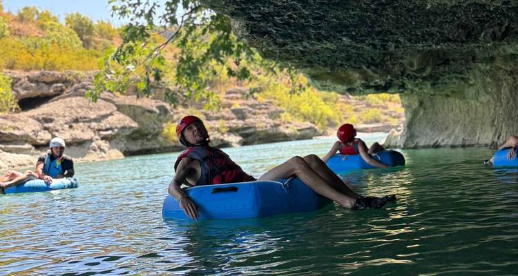 Travelers wearing helmets float on blue inner tubes along a calm river beneath an overhanging rock ledge and leafy branches.