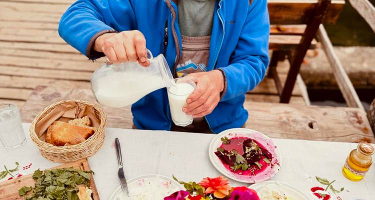 A person pours fresh milk from a glass jug into a tumbler during an outdoor rustic meal featuring homemade bread and salad.