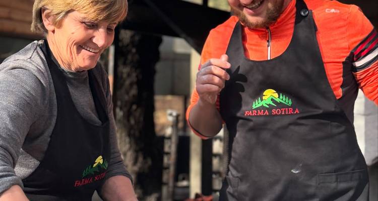 Two cooks wearing black aprons smile while preparing food together at an outdoor kitchen.