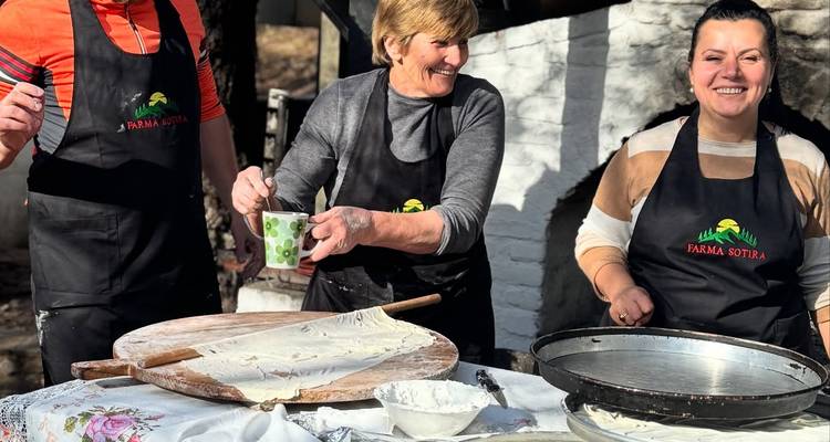 Three people stretch and roll out thin dough on a wooden table in a rustic outdoor kitchen, laughing together.