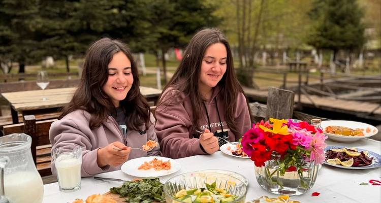 Two teenage girls enjoy a homemade outdoor meal at a long wooden table decorated with fresh flowers and local dishes.