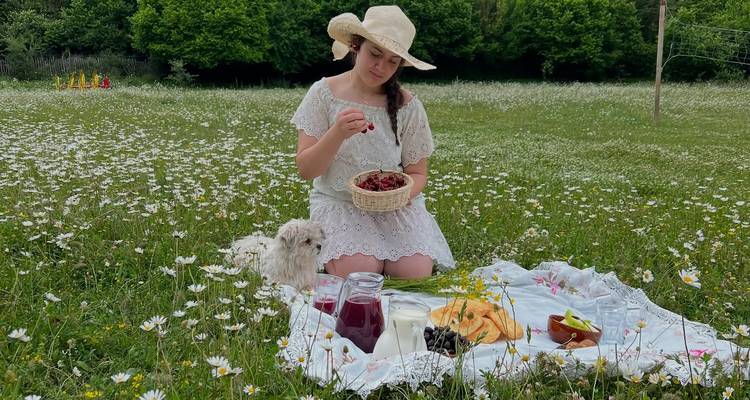 A young woman in a white dress prepares a picnic amid a field of daisies with a small white dog beside her.