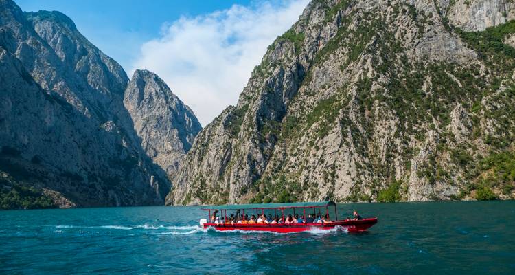 A red passenger boat filled with visitors cruises through the dramatic canyon walls of a turquoise lake.