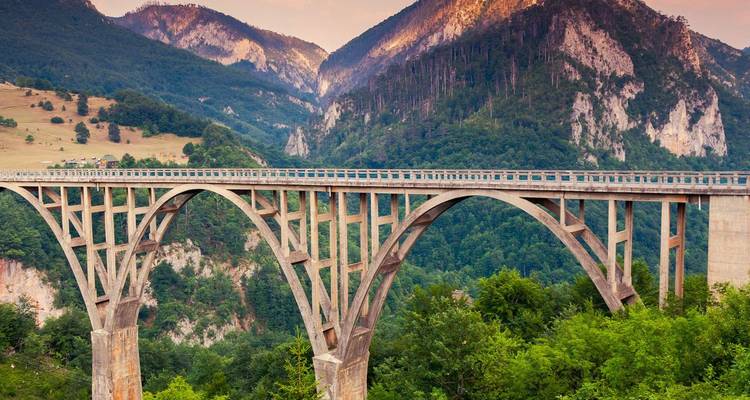 Die historische gewölbte Betonbrücke Tara überspannt eine tiefe bewaldete Schlucht vor der Kulisse schroffer Gipfel.