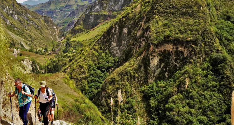 Excursionistas con bastones de trekking ascendiendo por un sendero rocoso estrecho entre paredes empinadas y verdes de un cañón en los Andes de Ecuador.