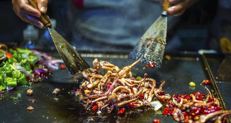 Close-up of a street food chef stir-frying spicy squid and vegetables on a sizzling griddle at night.