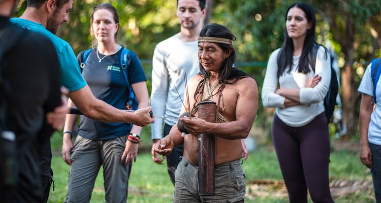 Los visitantes se reúnen alrededor de un guía indígena que sostiene instrumentos tradicionales en un claro sombreado del bosque.