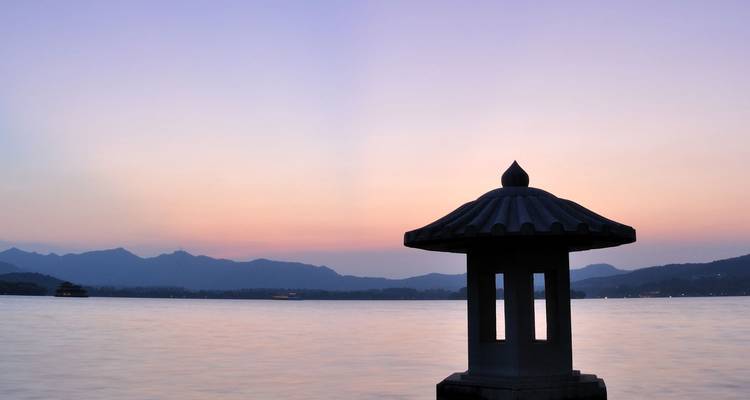 Silhouette of a stone lantern pavilion on West Lake at dusk with pastel sunset colours and distant mountains.