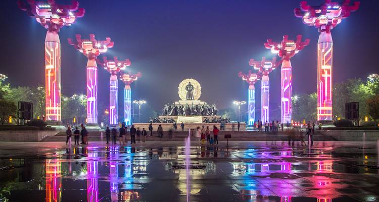 Colourful illuminated pillars and fountain reflections create a vibrant night scene in a city square.