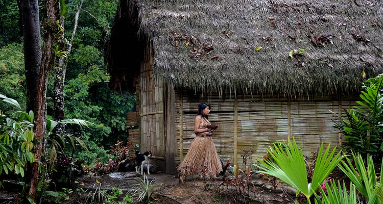 Inheemse vrouw in traditionele kledij die voorbij een rieten junglehut loopt met dichte begroeiing.