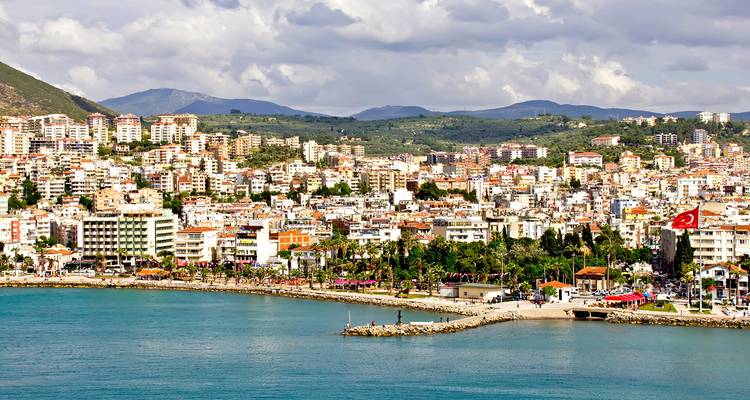 Seaside skyline of Kusadasi backed by green hills under a partly cloudy sky.