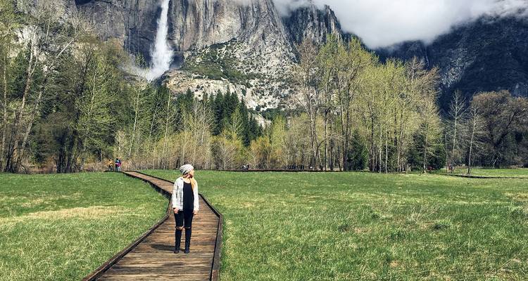 Viajero caminando por un sendero de madera a través de un prado con hierba hacia un acantilado de granito con cascadas en Yosemite.