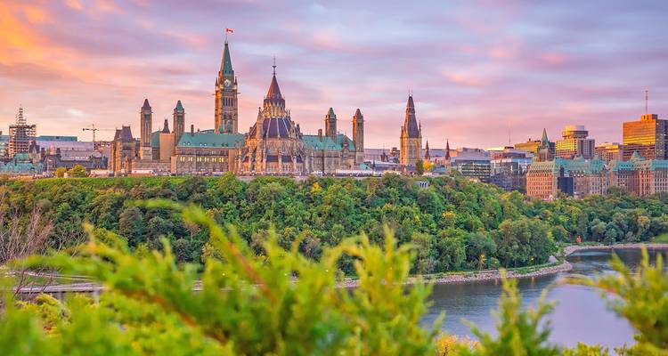 Sunset light bathes Ottawa’s Parliament Buildings perched above the Ottawa River with lush foreground greenery.