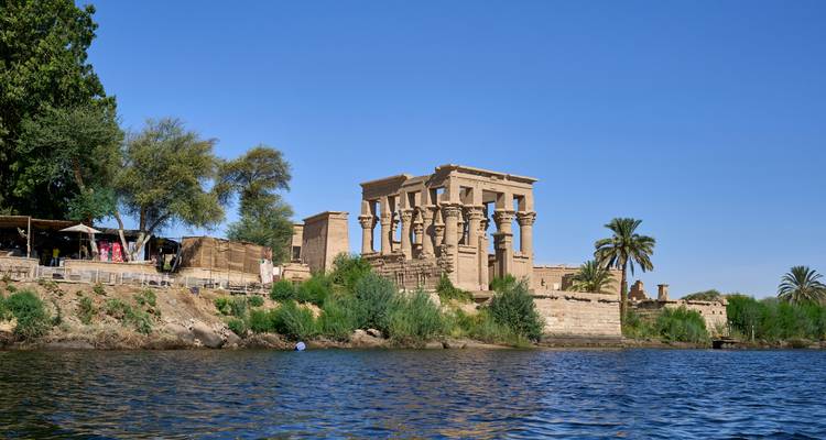 Ancient stone temple ruins standing on an island along a calm Nile River under a clear blue sky.
