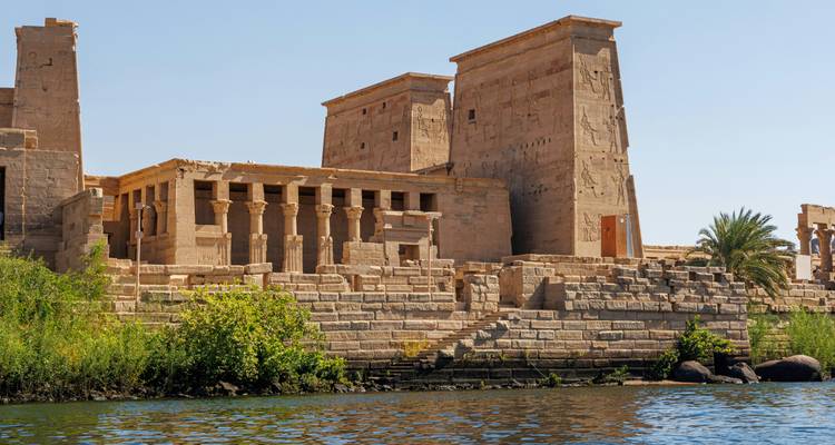 Sand-colored temple walls and columns rise directly from the Nile’s edge on a bright day.