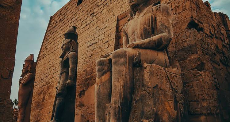 Massive seated and standing pharaonic statues carved from stone at an ancient temple entrance.