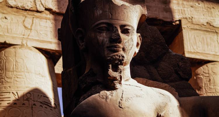 Close-up of a weathered pharaoh statue head illuminated by warm sunlight among temple pillars.