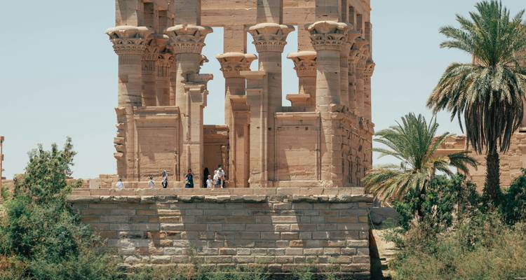Visitors stand on a stone terrace of an Egyptian riverside temple surrounded by green shrubs.
