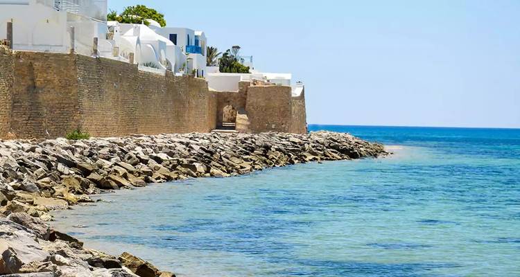 Zonnig kustgezicht van een stenen zeewering omzoomd met witgekalkte mediterrane huizen naast helder turquoise water.