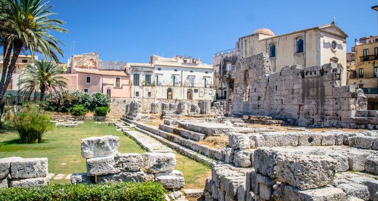 Ruins of ancient stone temple columns and walls stand beside Mediterranean palm trees under bright blue sky.
