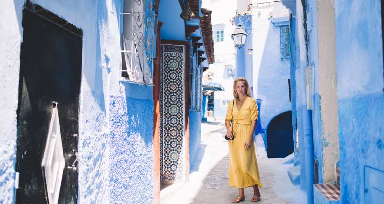 Mujer en un vestido amarillo brillante se encuentra en un callejón azul estrecho de Chefchaouen iluminado por suave luz del día.