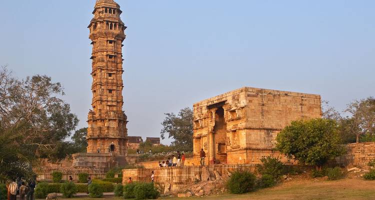 Der Siegesturm des Forts Chittorgarh erhebt sich neben alten Steintempeln und spazierenden Besuchern auf grasbewachsenen Flächen.
