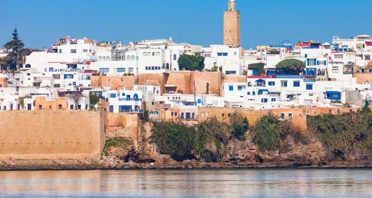 Fortificaciones ribereñas y casas blanco-azules de Rabat reflejadas en aguas tranquilas en un día soleado.