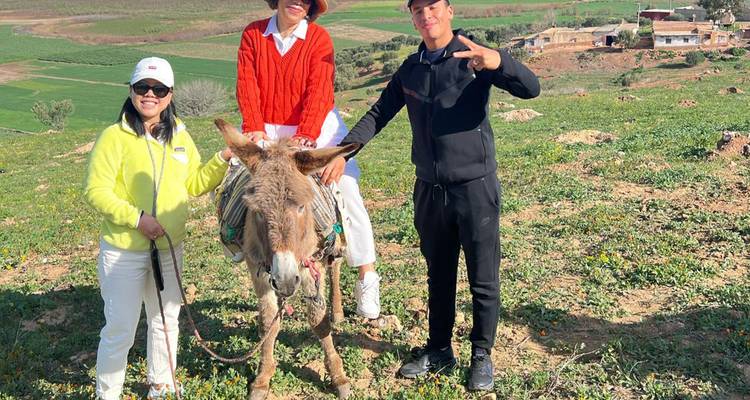 Trois voyageurs sourient à côté d'un âne dans un paysage rural et verdoyant du Maroc.