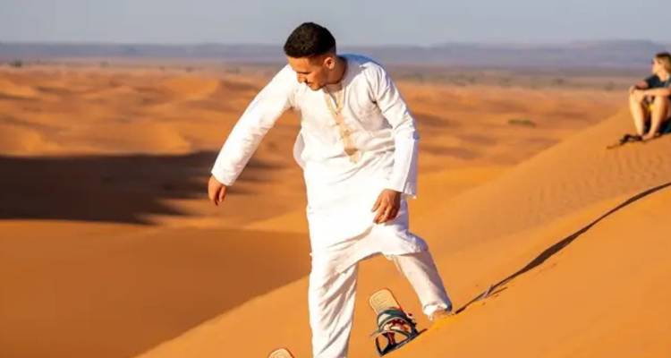 Man in traditional robe sandboarding down orange Sahara dune at sunset.