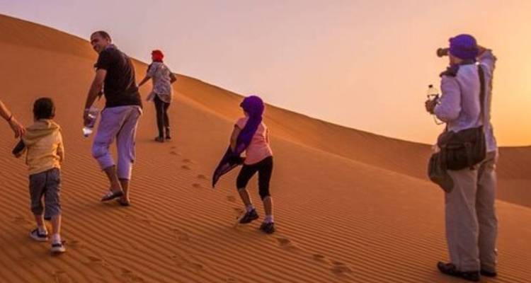 Family trekking up a sand dune at dusk leaving footprints under warm orange light.