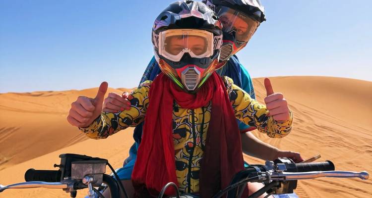Couple wearing helmets gives thumbs-up while riding a quad bike across Sahara dunes.