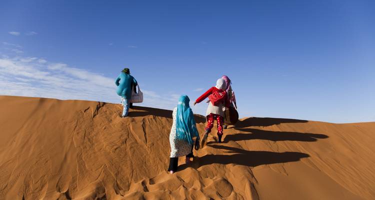 Four travellers in colourful scarves trek up a golden sand dune under clear sky.