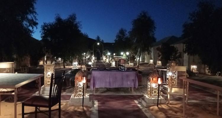 Nighttime desert camp dining area lit by lanterns with tables set under starry sky.