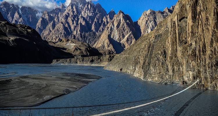 Puente colgante peatonal se extiende sobre un río glacial en medio de imponentes montañas escarpadas bajo un cielo azul brillante.