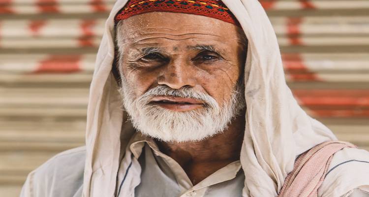 Retrato cercano de un hombre mayor con barba que lleva una gorra con diseños y un chal claro.