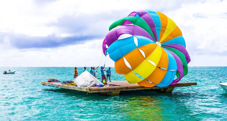 Crew prepares a colourful rainbow parasail on a floating platform over clear turquoise lagoon water.