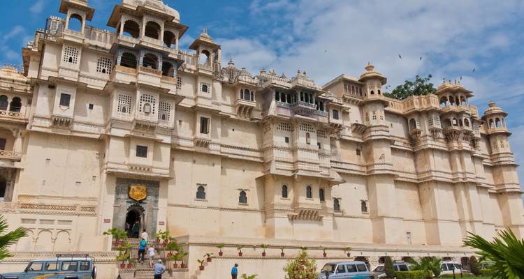 Große weiße Steinpalast-Fassade unter einem hellen Himmel mit Besuchern und geparkten Autos davor.