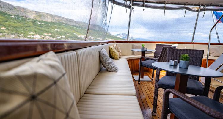 Cozy cushioned bench and small tables on a shaded deck of a yacht overlooking green hills and sea.