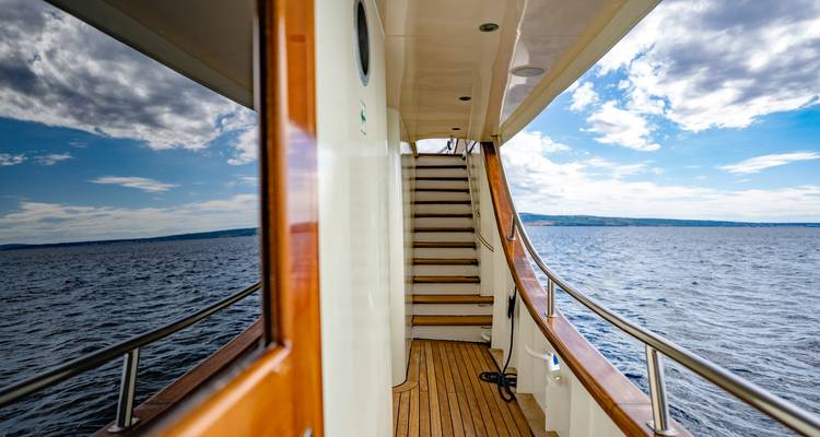 Side corridor of a yacht with wooden stairs leading upward and ocean views reflected on the window.