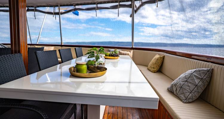 Outdoor dining table and cushioned benches on covered yacht deck with ocean backdrop.