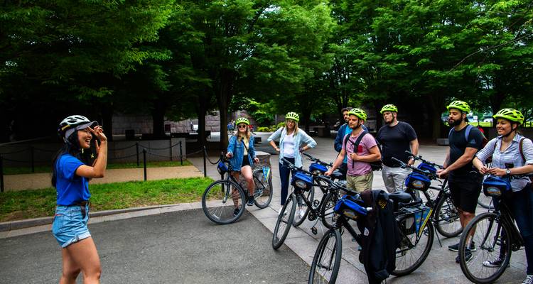 Reiseführer, der eine kleine Gruppe von Radfahrern mit Helmen in einer grünen städtischen Parkumgebung anspricht.