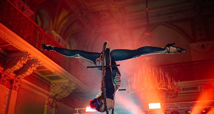 Un acrobate exécute un grand écart à l'envers sur un agrès aérien sous un éclairage de scène dramatique.