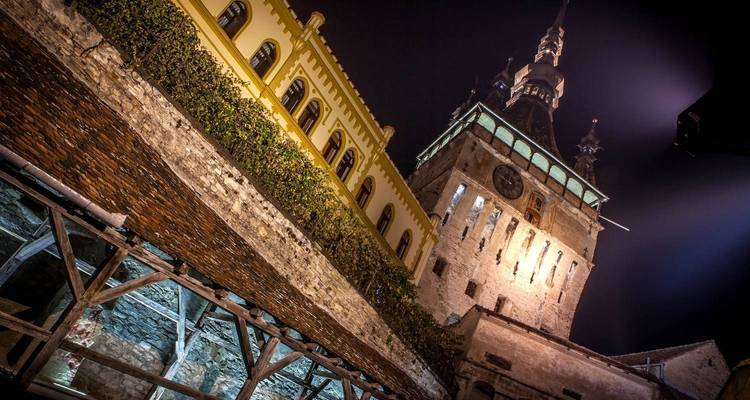 Vue nocturne de la Tour de l'Horloge de Sighișoara et de l'escalier couvert avec un éclairage dramatique.