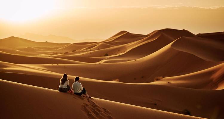 Paar sitzt auf goldenen Sanddünen und beobachtet den Sonnenuntergang über hügeligen Wüstenlandschaften.