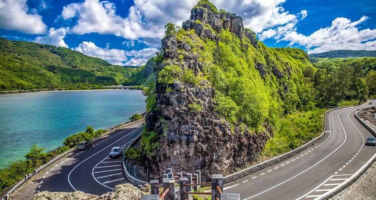 Dramatic coastal road curving around a rocky green promontory beside turquoise lagoon under bright sky