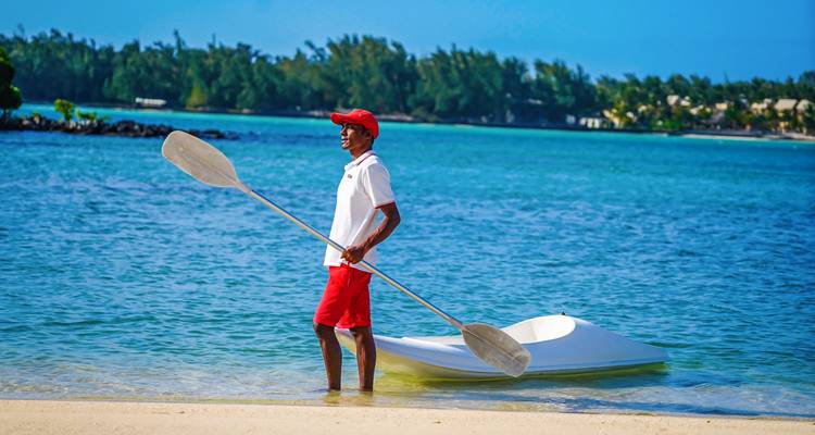 Socorrista de playa con gorra roja y pantalones cortos sosteniendo un remo junto a una laguna cristalina y un bote pequeño