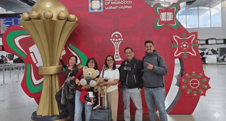 Des voyageurs souriant devant un présentoir d'aéroport présentant une maquette géante du trophée de la Coupe d'Afrique.